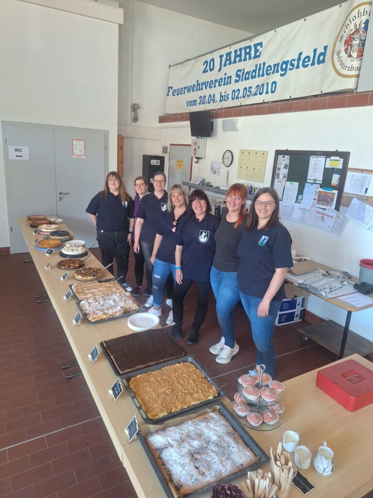 Sieben Frauen stehen in einem hellen Raum hinter einem langen Tisch mit einer Vielzahl von Kuchen und Gebäck. Darüber hängt ein Banner, mit dem das 20-jährige Bestehen des örtlichen Feuerwehrvereins gefeiert wird.