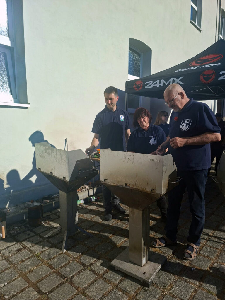 Drei Personen in dunklen Hemden stehen an zwei Metallgrills auf einem sonnigen, gepflasterten Außengelände, neben einem hellen Gebäude und einem schwarzen Vordach mit 24MX-Logos.