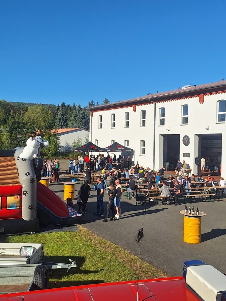 Die Menschen versammeln sich vor einem weißen Gebäude mit roter Umrandung, sitzen an Picknicktischen und stehen in Gruppen. Es gibt eine aufblasbare Rutsche in Form eines Hundes und Getränkeflaschen auf Fässern unter einem klaren blauen Himmel.