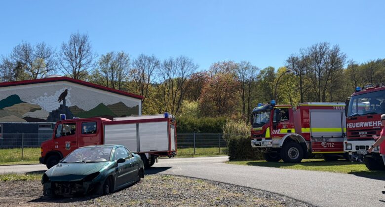 Ein beschädigtes grünes Auto steht neben einer Straße, während zwei Feuerwehrfahrzeuge in der Nähe geparkt sind. Feuerwehrleute stehen in der Nähe der Fahrzeuge, und im Hintergrund sind ein Gebäude mit einer Wandmalerei und Bäume unter einem klaren blauen Himmel zu sehen.
