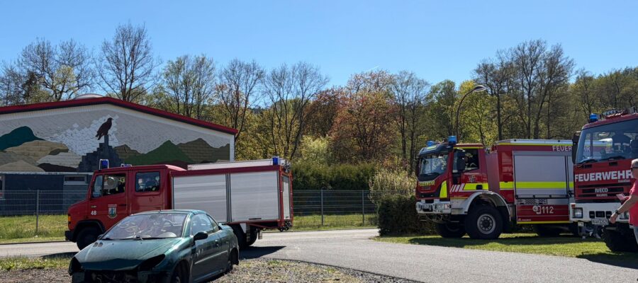 Ein beschädigtes grünes Auto steht neben einer Straße, während zwei Feuerwehrfahrzeuge in der Nähe geparkt sind. Feuerwehrleute stehen in der Nähe der Fahrzeuge, und im Hintergrund sind ein Gebäude mit einer Wandmalerei und Bäume unter einem klaren blauen Himmel zu sehen.