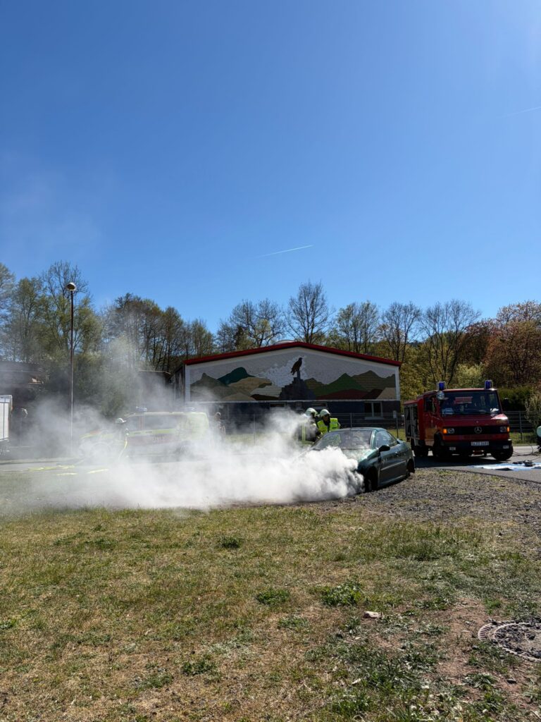 Feuerwehrleute löschen an einem sonnigen Tag den Rauch aus einem Auto auf einer offenen Wiese mit einem Feuerwehrauto in der Nähe und einem Gebäude mit einer Wandmalerei im Hintergrund.