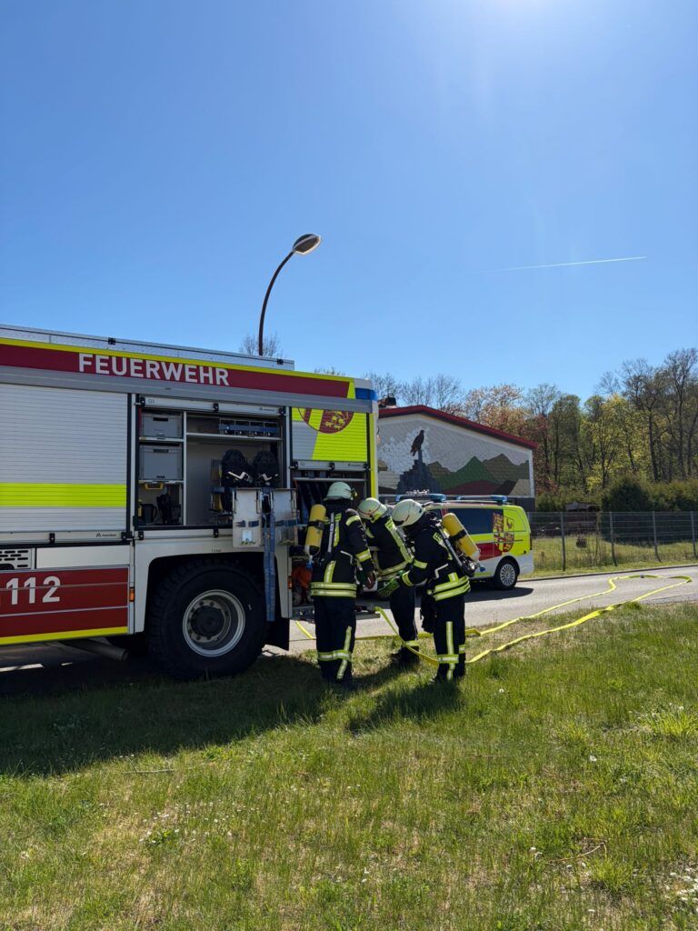 Drei Feuerwehrmänner in voller Montur stehen an einem sonnigen Tag neben einem Feuerwehrauto mit der Aufschrift "Feuerwehr", mit Schläuchen auf dem Boden und einem Wandbild mit Bergen und einer Person im Hintergrund.