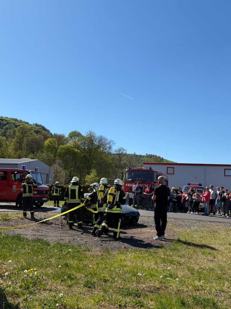 Feuerwehrmänner in voller Montur verwenden Geräte in der Nähe eines Autos, während eine Menschenmenge zusieht. Feuerwehrfahrzeuge sind in der Nähe geparkt, und die Szene spielt an einem sonnigen Tag im Freien mit klarem Himmel und grünen Bäumen im Hintergrund.