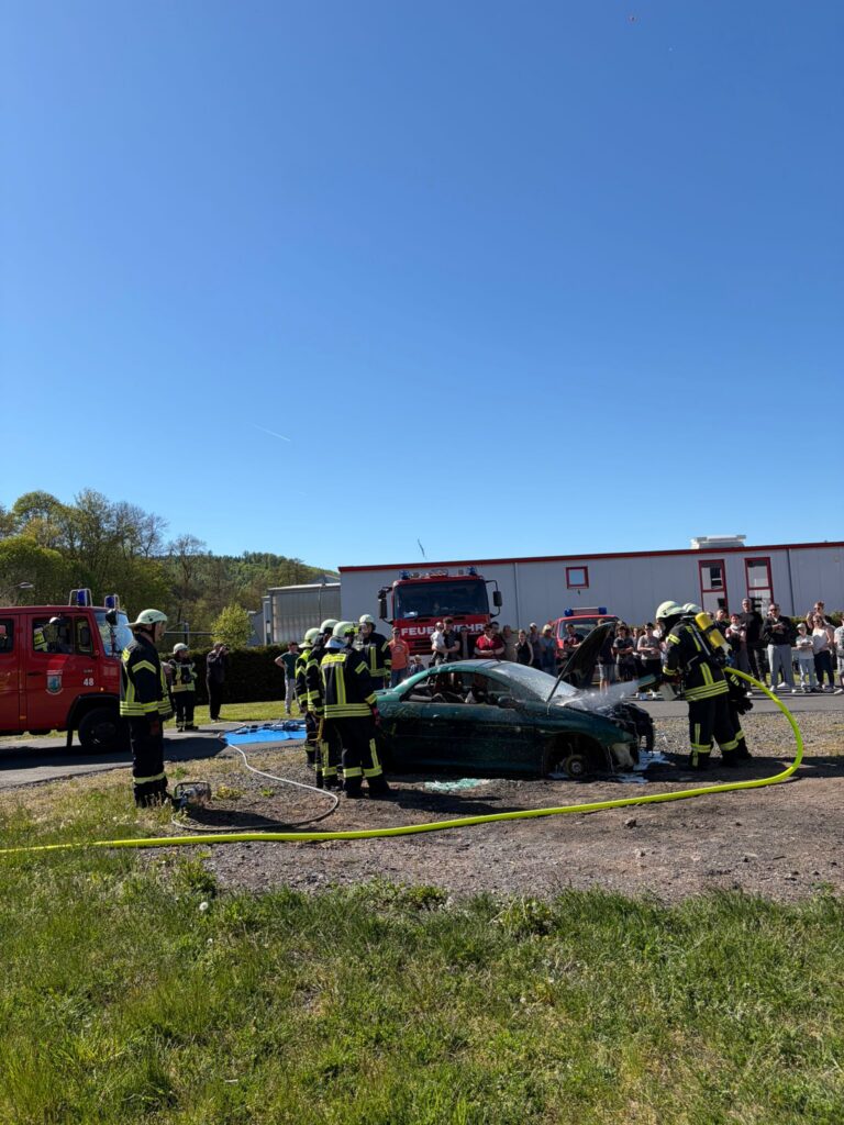 Feuerwehrleute in voller Montur umringen ein schwer beschädigtes Auto während einer Rettungsdemonstration. Im Hintergrund sind bei strahlend blauem Himmel mehrere Feuerwehrfahrzeuge und eine Gruppe von Schaulustigen zu sehen.