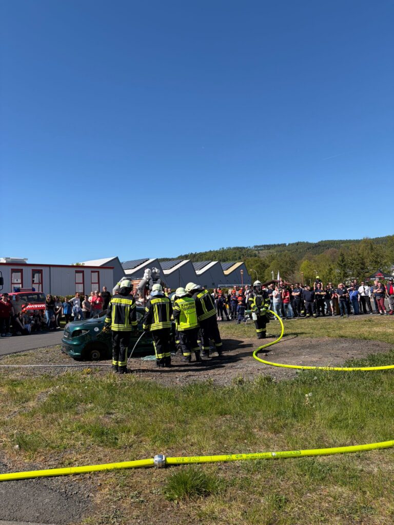 Feuerwehrleute in gelber und schwarzer Ausrüstung führen eine Vorführung um ein grünes Auto herum durch, während eine große Menschenmenge an einem sonnigen Tag im Freien zusieht, mit Gebäuden und Bäumen im Hintergrund.