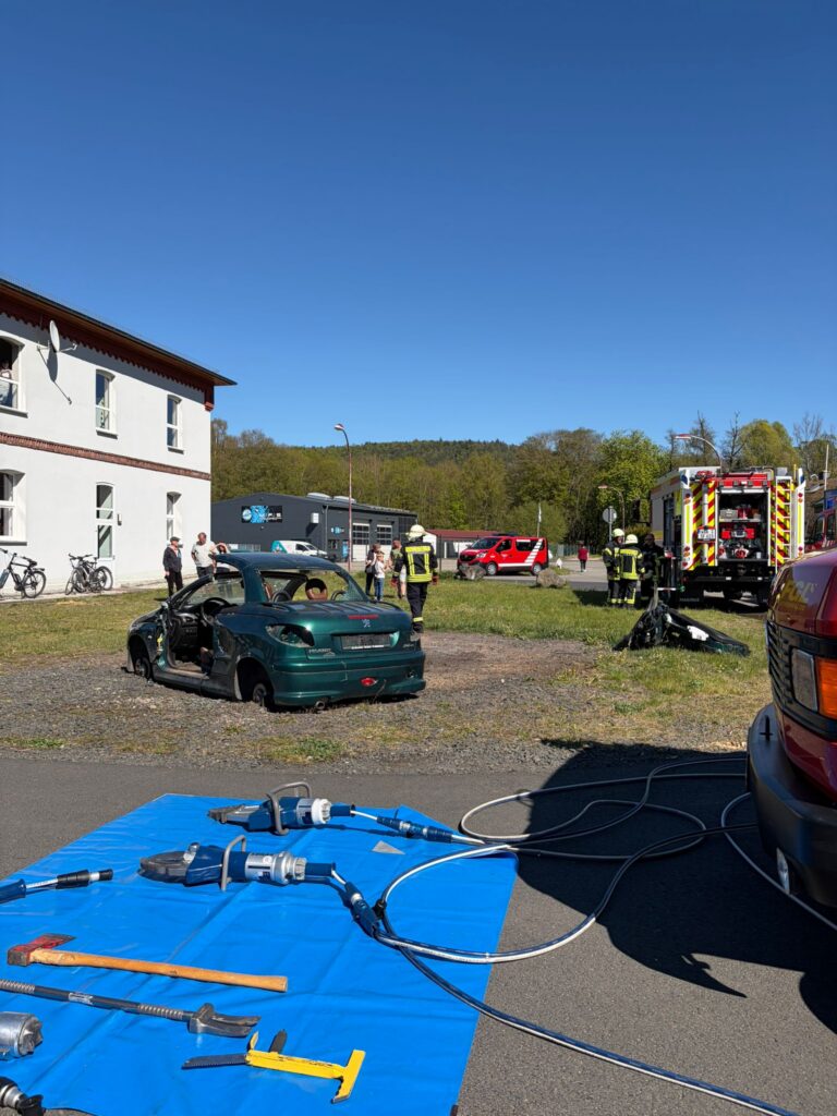 Ein grünes Auto mit abgenommenem Dach steht neben einem Gebäude. Im Hintergrund sind Feuerwehrleute und Einsatzfahrzeuge zu sehen. Rettungsgeräte und Schläuche sind auf einer blauen Plane im Vordergrund ausgelegt. Bei der Szene scheint es sich um eine Übung zu handeln.