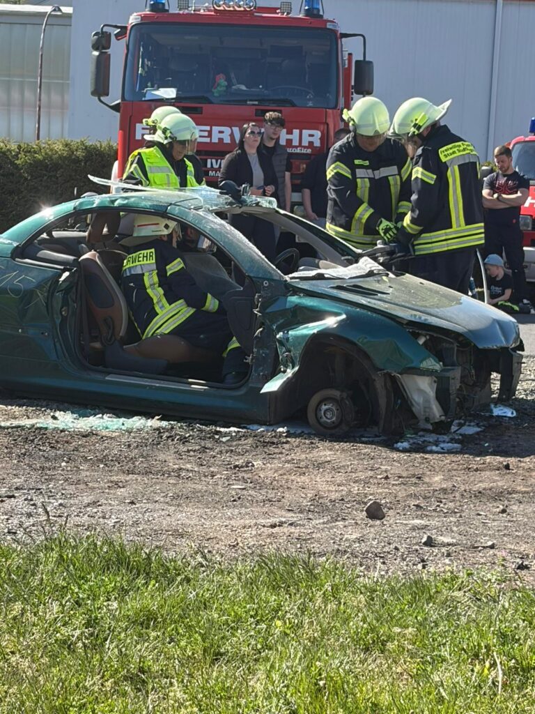 Feuerwehrleute in voller Montur arbeiten an der Stelle eines Autounfalls, an der einem Autowrack ein Vorderrad fehlt. Ein Feuerwehrauto und mehrere Schaulustige sind an einem sonnigen Tag im Hintergrund zu sehen.