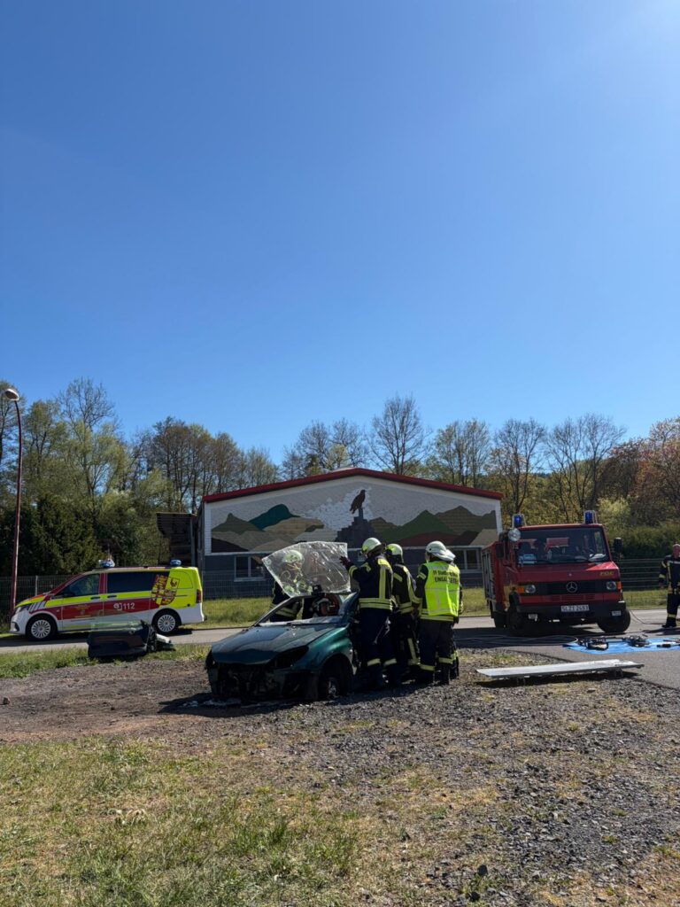 Mehrere Feuerwehrleute in Schutzkleidung stehen um ein beschädigtes Auto herum, während in der Nähe Einsatzfahrzeuge geparkt sind. Im Hintergrund ist ein Gebäude mit einer Wandmalerei und Bäumen unter einem strahlend blauen Himmel zu sehen.