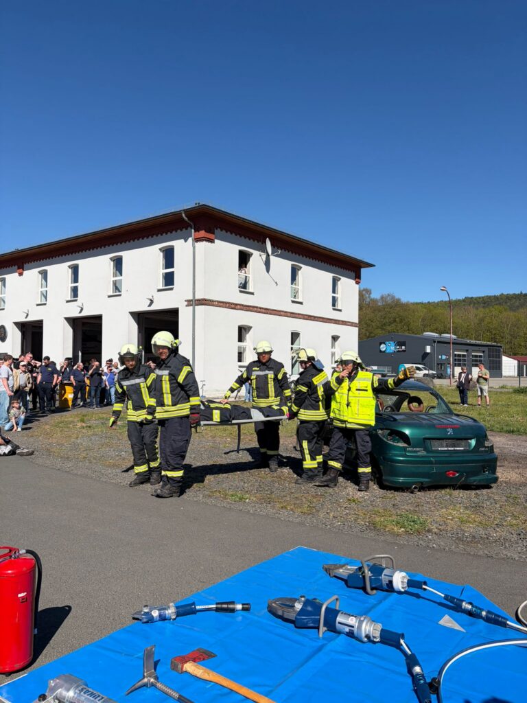 Feuerwehrleute in voller Montur tragen eine Trage während einer Rettungsdemonstration neben einem beschädigten grünen Auto, während eine Menschenmenge in der Nähe eines weißen Gebäudes zuschaut und im Vordergrund Notfallausrüstung zu sehen ist.