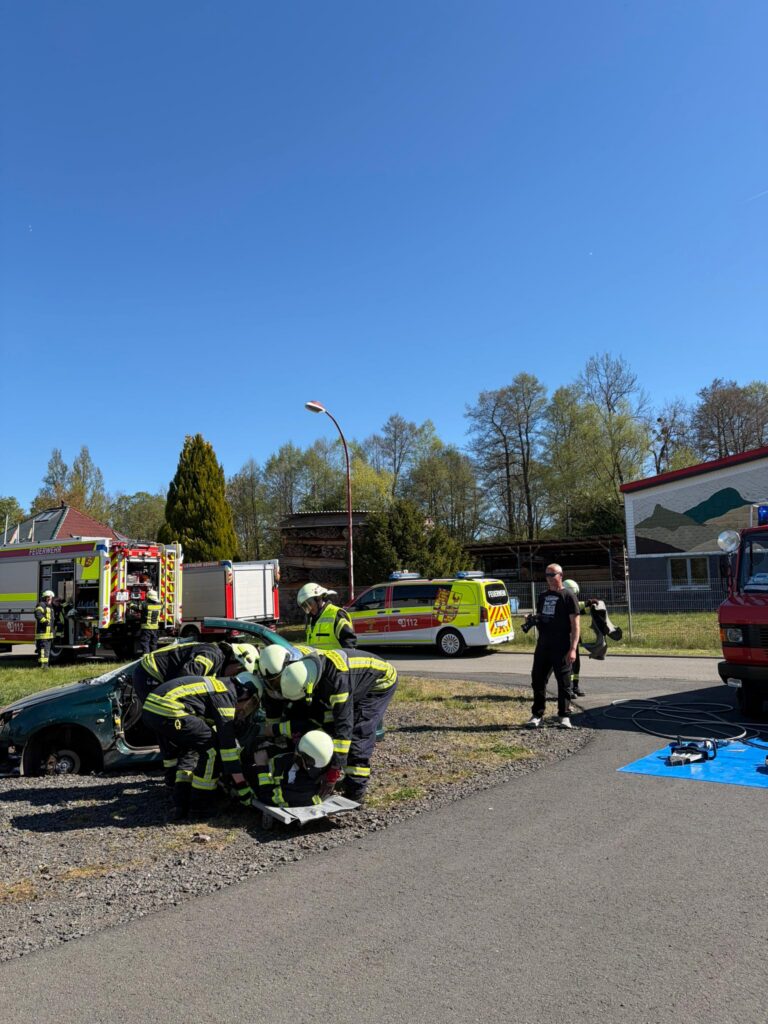 Mehrere Feuerwehrleute in voller Montur arbeiten gemeinsam neben einem beschädigten grünen Auto, während Einsatzfahrzeuge und einige Menschen an einem sonnigen Tag mit klarem blauen Himmel und Bäumen im Hintergrund in der Nähe stehen.