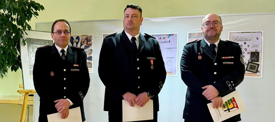 Drei Männer in formellen Uniformen stehen in einem Innenraum nebeneinander und halten jeweils eine Urkunde in der Hand. Sie scheinen an einer Zeremonie teilzunehmen, im Hintergrund sind Plakate und eine Pflanze zu sehen.