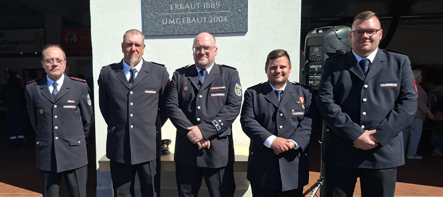 Fünf Männer in formellen dunklen Uniformen stehen nebeneinander im Freien im Sonnenlicht und posieren vor einer weißen Wand mit einer Gedenktafel. Sie scheinen zu einer Feuerwehr oder einem Rettungsdienst zu gehören.