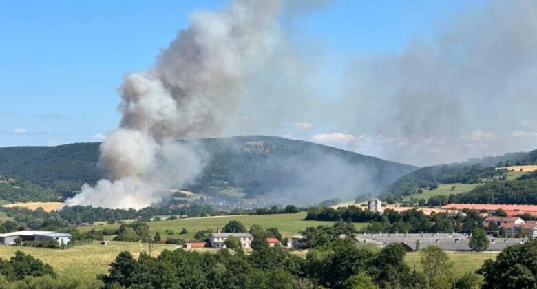 Dichter Rauch steigt von einem Waldbrand in einem ländlichen Tal auf, im Vordergrund sind Hügel und Häuser vor blauem Himmel zu sehen.