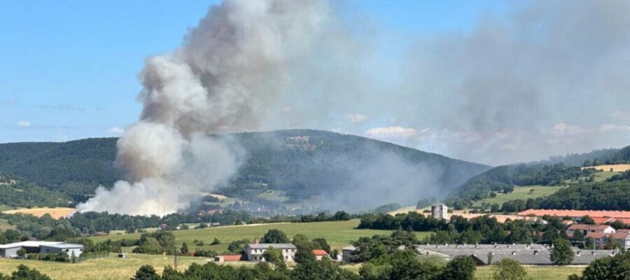 Dichter Rauch steigt von einem Waldbrand in einem ländlichen Tal auf, im Vordergrund sind Hügel und Häuser vor blauem Himmel zu sehen.
