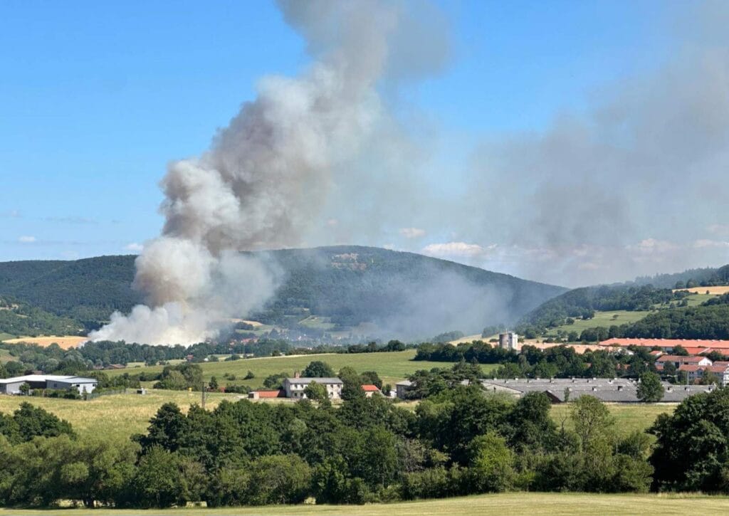 Dichter Rauch steigt von einem Waldbrand in einem ländlichen Tal auf, im Vordergrund sind Hügel und Häuser vor blauem Himmel zu sehen.