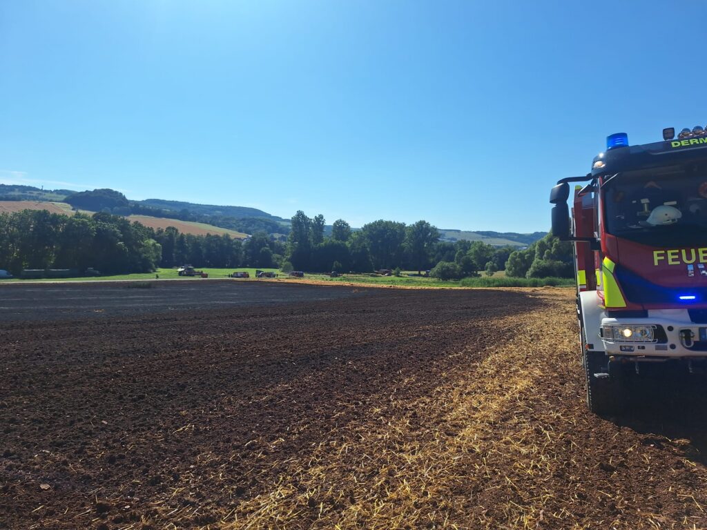 Unter einem strahlend blauen Himmel steht ein Feuerwehrauto am Rande eines großen, frisch gepflügten Feldes, im Hintergrund sind Bäume und Hügel zu sehen.