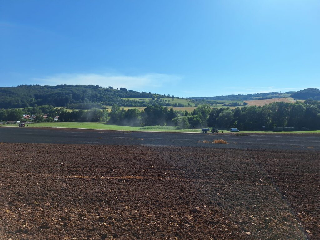Im Vordergrund ein gepflügtes Feld, im Hintergrund grüne Hügel, Bäume und vereinzelte Häuser unter einem strahlend blauen Himmel an einem sonnigen Tag.
