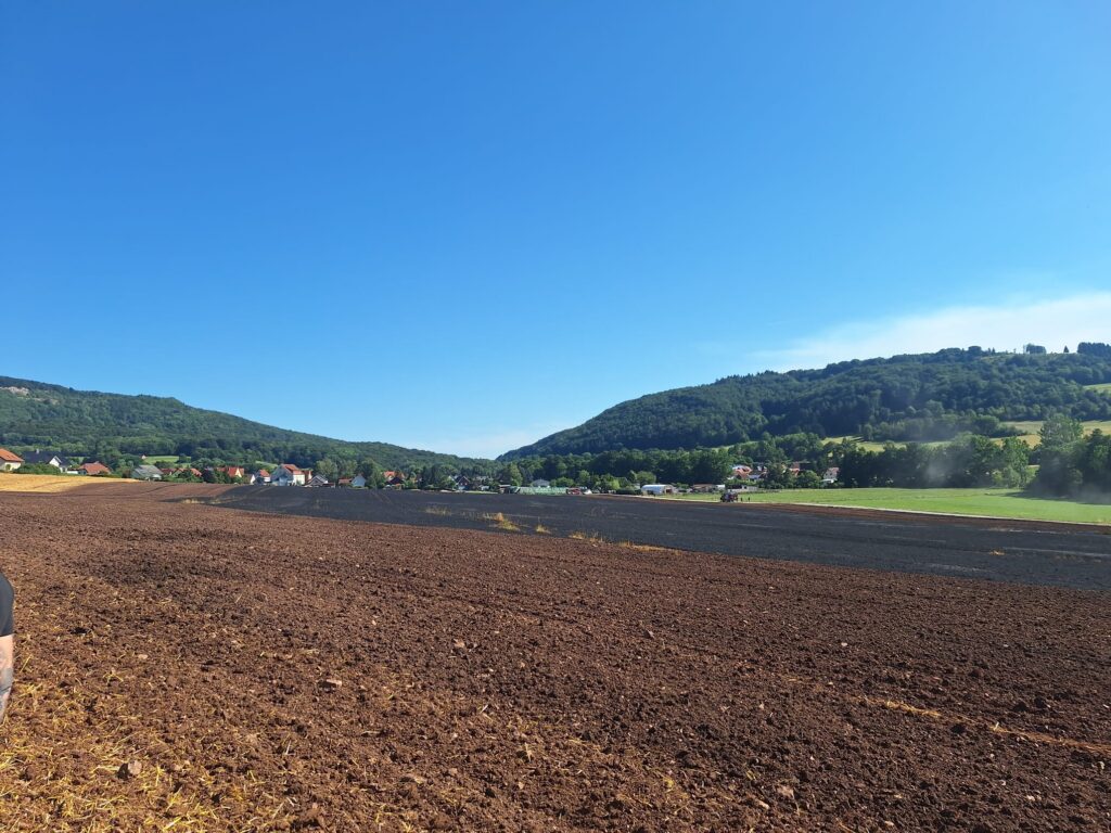 Ein frisch gepflügtes Feld unter strahlend blauem Himmel, im Hintergrund grüne Hügel und ein kleines Dorf mit bunten Häusern.