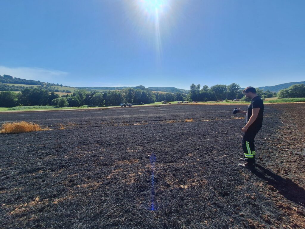 Eine Person in Schutzkleidung steht unter strahlendem Sonnenschein auf einem großen, verbrannten Feld, im Hintergrund sind grüne Bäume und Hügel zu sehen.