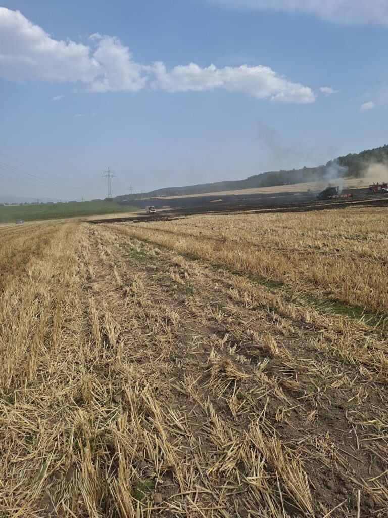 Ein trockenes, abgeerntetes Feld, über dem in der Ferne Rauch aufsteigt, möglicherweise von einem Feuer. Der Himmel ist größtenteils klar mit wenigen Wolken, und Stromleitungen ziehen sich über den Horizont. In der Nähe des Rauchs sind einige Maschinen zu sehen.