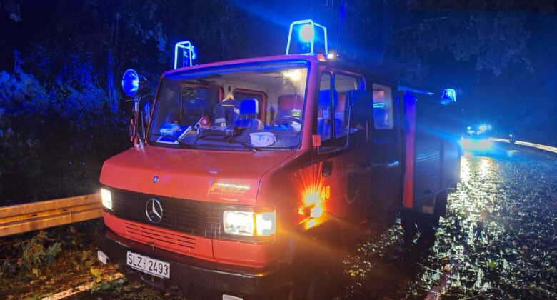 Ein rotes Feuerwehrauto mit blinkenden blauen Lichtern steht nachts auf einer nassen, mit Laub bedeckten Straße. Überall liegen Trümmer und Äste verstreut, und im Hintergrund sind weitere Warnleuchten zu sehen.