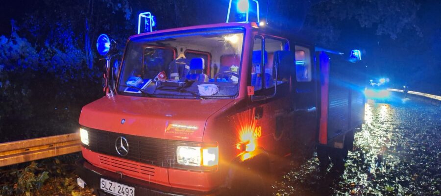 Ein rotes Feuerwehrauto mit blinkenden blauen Lichtern steht nachts auf einer nassen, mit Laub bedeckten Straße. Überall liegen Trümmer und Äste verstreut, und im Hintergrund sind weitere Warnleuchten zu sehen.