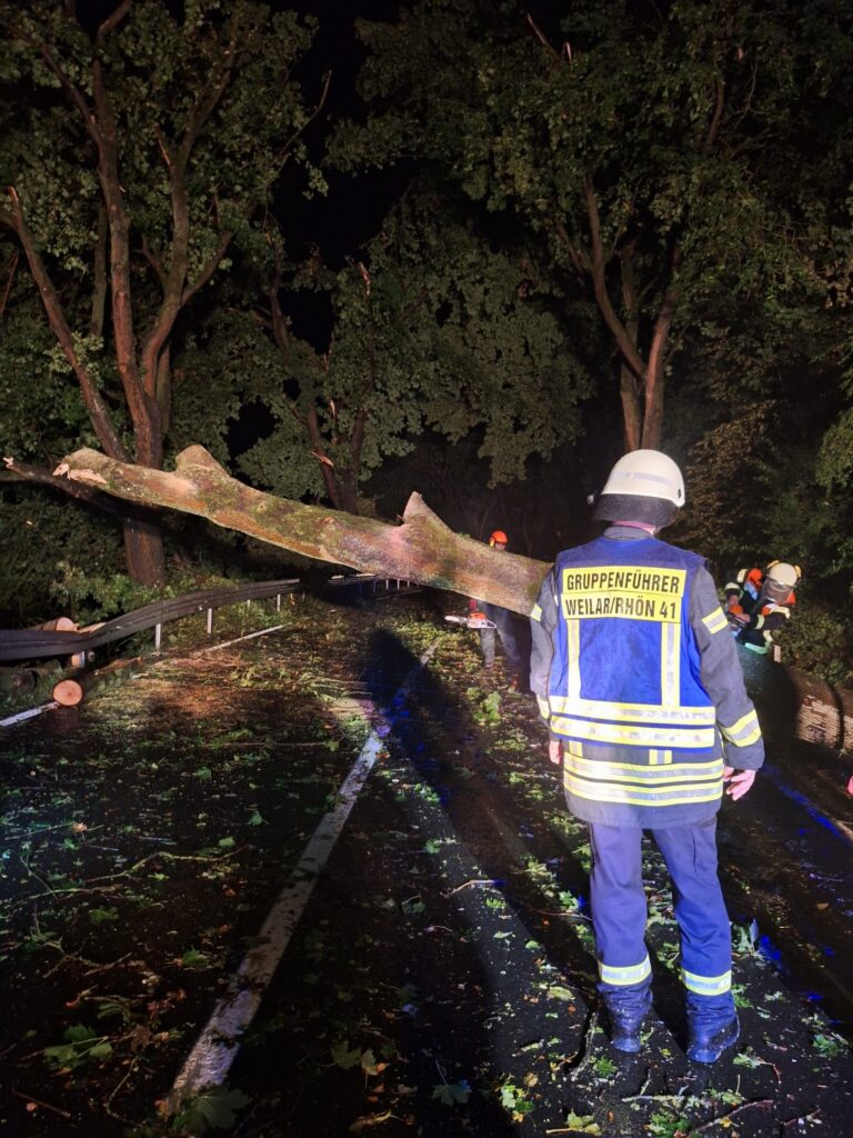 Feuerwehrleute arbeiten nachts daran, einen umgestürzten Baum zu beseitigen, der eine Straße blockiert. Im Vordergrund steht ein Feuerwehrmann in Uniform, im Hintergrund sind weitere Feuerwehrleute zu sehen, die mit Werkzeugen arbeiten. Trümmer und Laub bedecken die nasse Straße.