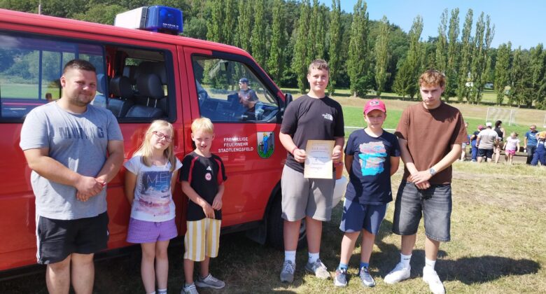 Sechs Personen, darunter Kinder und Erwachsene, stehen an einem sonnigen Tag auf einer offenen Wiese neben einem roten Rettungsfahrzeug. Eine Person hält eine Urkunde in der Hand. Im Hintergrund sind Bäume und Menschen zu sehen.