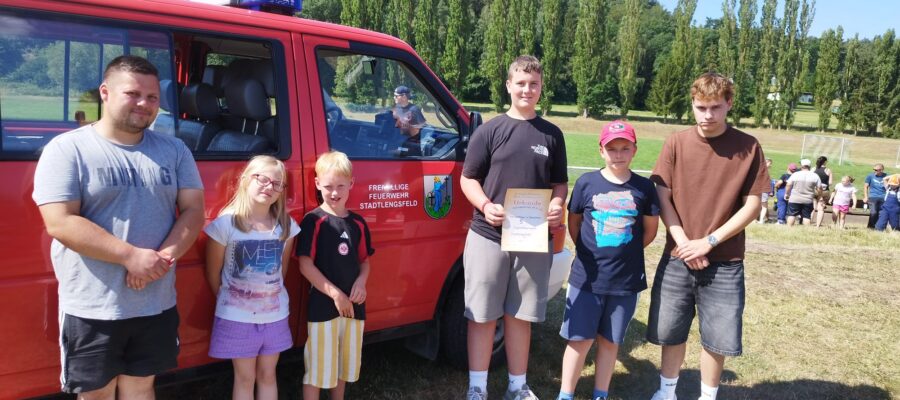 Sechs Personen, darunter Kinder und Erwachsene, stehen an einem sonnigen Tag auf einer offenen Wiese neben einem roten Rettungsfahrzeug. Eine Person hält eine Urkunde in der Hand. Im Hintergrund sind Bäume und Menschen zu sehen.