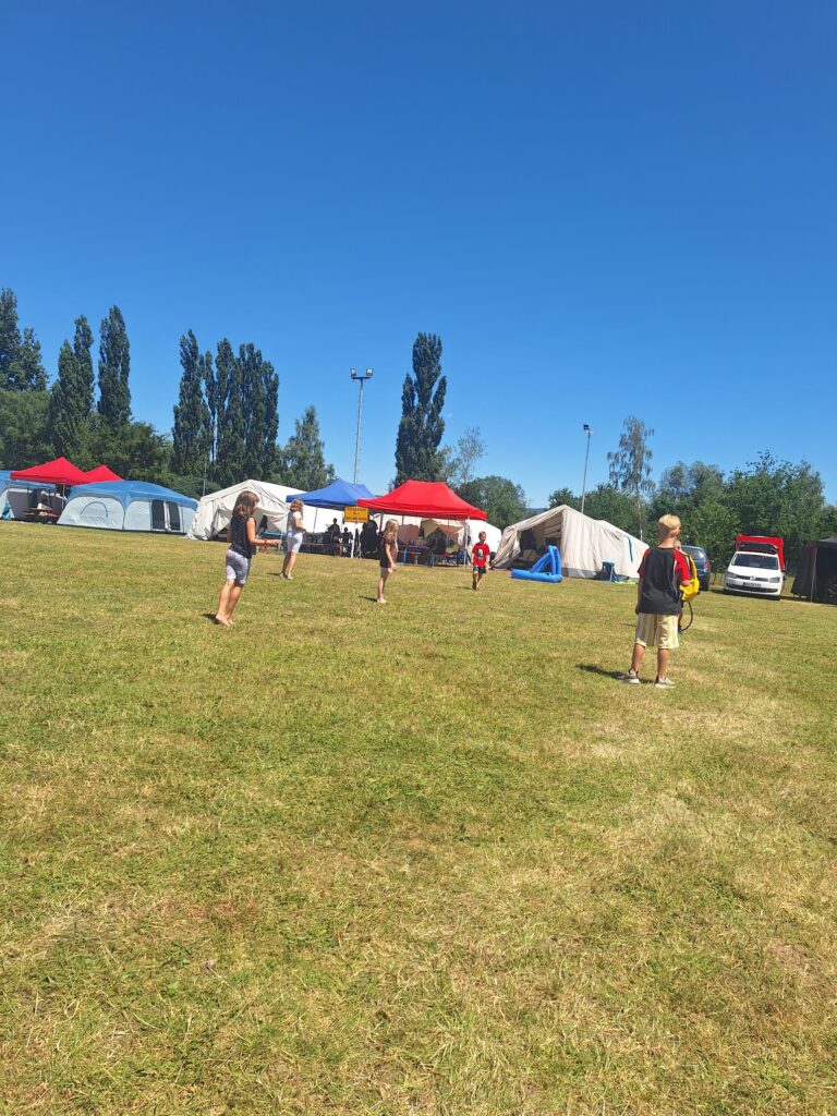 Kinder spielen auf einer Wiese mit Zelten, einem roten Baldachin und Bäumen im Hintergrund unter strahlend blauem Himmel. Einige Erwachsene stehen in der Nähe der Zelte, und rechts ist ein weißer Transporter geparkt.