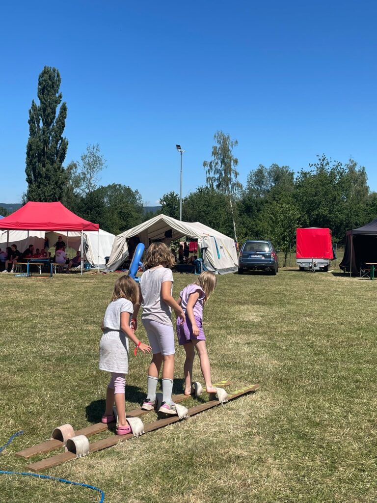 Drei Kinder balancieren im Freien auf Holzbrettern und Zylindern, im Hintergrund sind an einem sonnigen Tag Zelte, Menschen und ein Auto zu sehen.