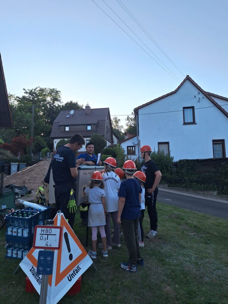 Eine Gruppe von Kindern mit orangefarbenen Helmen schaut zu, wie zwei Erwachsene im Freien in der Nähe von Häusern die Ausrüstung vorführen. Auf dem Rasen sind Sicherheitsschilder und Ausrüstungsgegenstände zu sehen.
