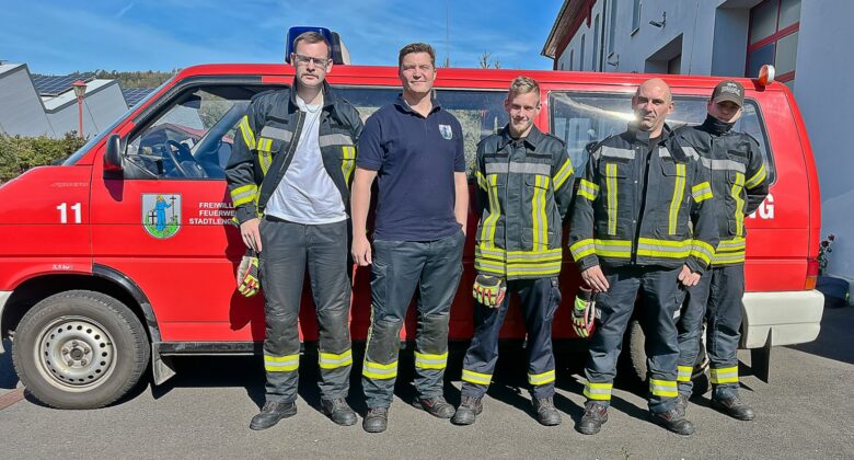 Fünf Feuerwehrleute stehen vor einem roten Feuerwehrauto, vier davon tragen Feuerwehruniform, einer ein marineblaues Hemd. Die Sonne scheint, und im Hintergrund sind Gebäude und ein strahlend blauer Himmel zu sehen.