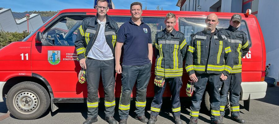 Fünf Feuerwehrleute stehen vor einem roten Feuerwehrauto, vier davon tragen Feuerwehruniform, einer ein marineblaues Hemd. Die Sonne scheint, und im Hintergrund sind Gebäude und ein strahlend blauer Himmel zu sehen.